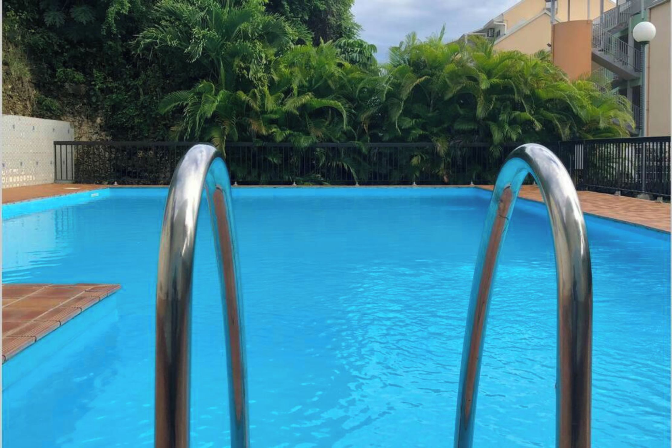 A swimming pool with metal ladders, surrounded by lush greenery and a building in the background.