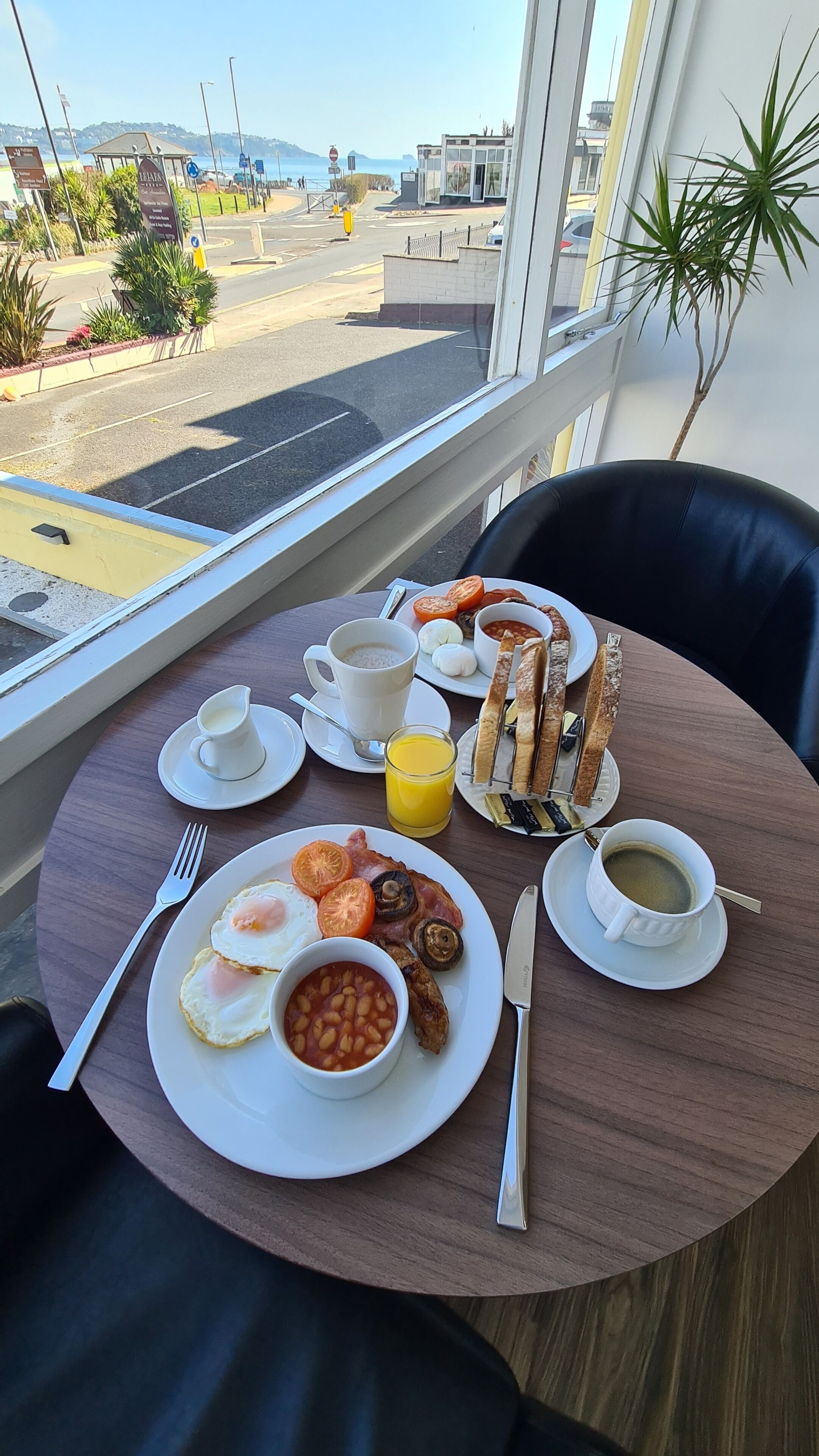 Ein Frühstückstisch mit Eiern, Bohnen, Toast und Saft, einer Tasse Kaffee und Blick auf das Meer und die Gebäude.