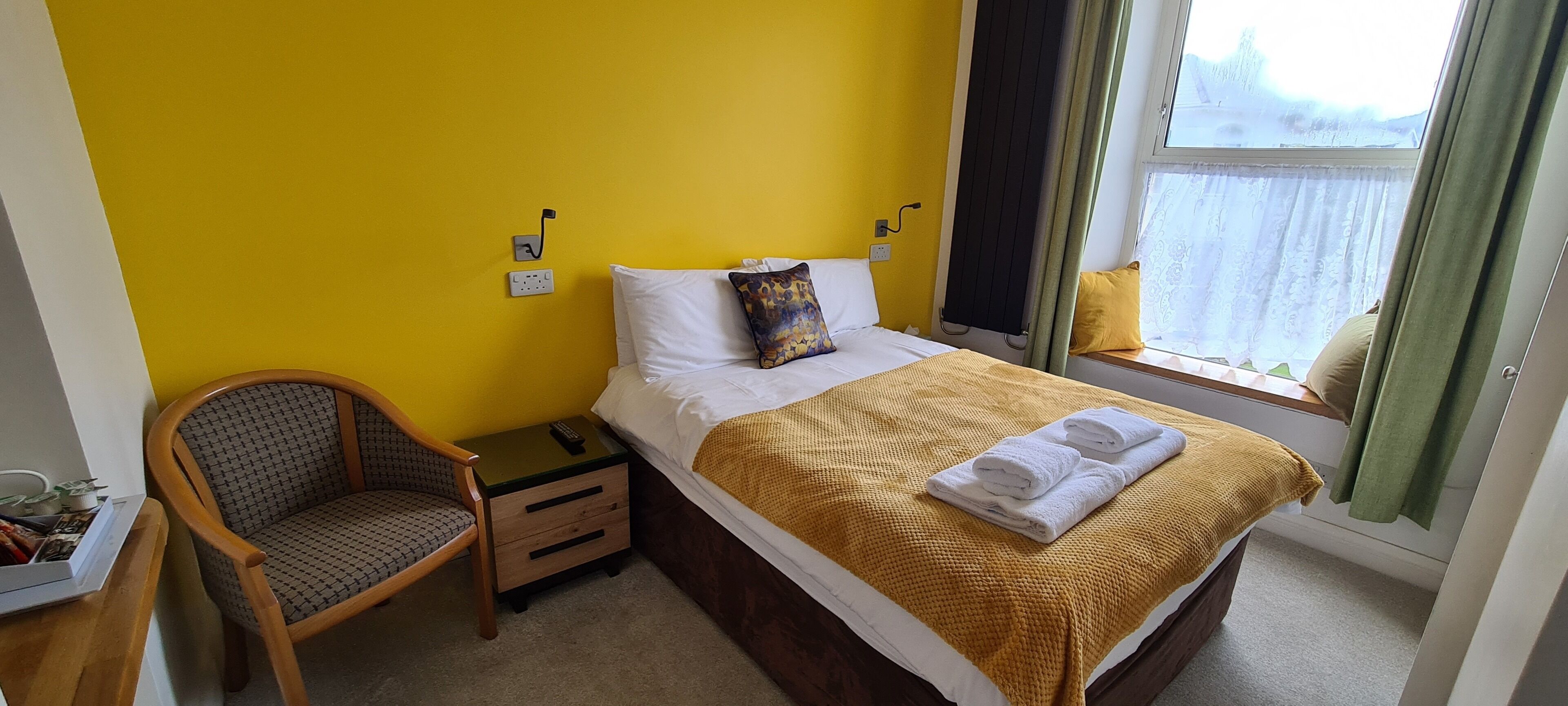 Une chambre d’hôtel avec un mur jaune, un lit recouvert d’une couverture jaune, une chaise, une table de chevet et une fenêtre avec des rideaux.