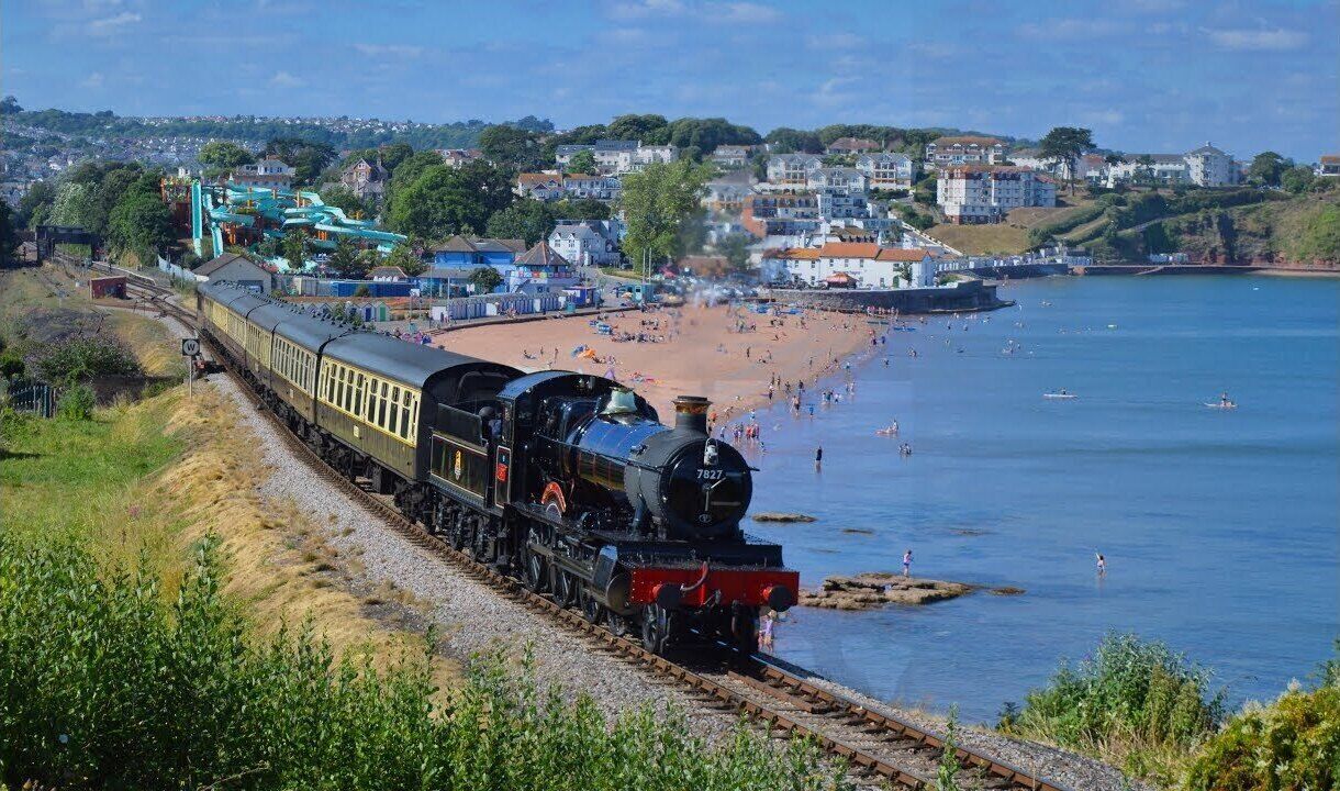 A train with a black engine and yellow carriages on tracks beside a beach with people and boats.