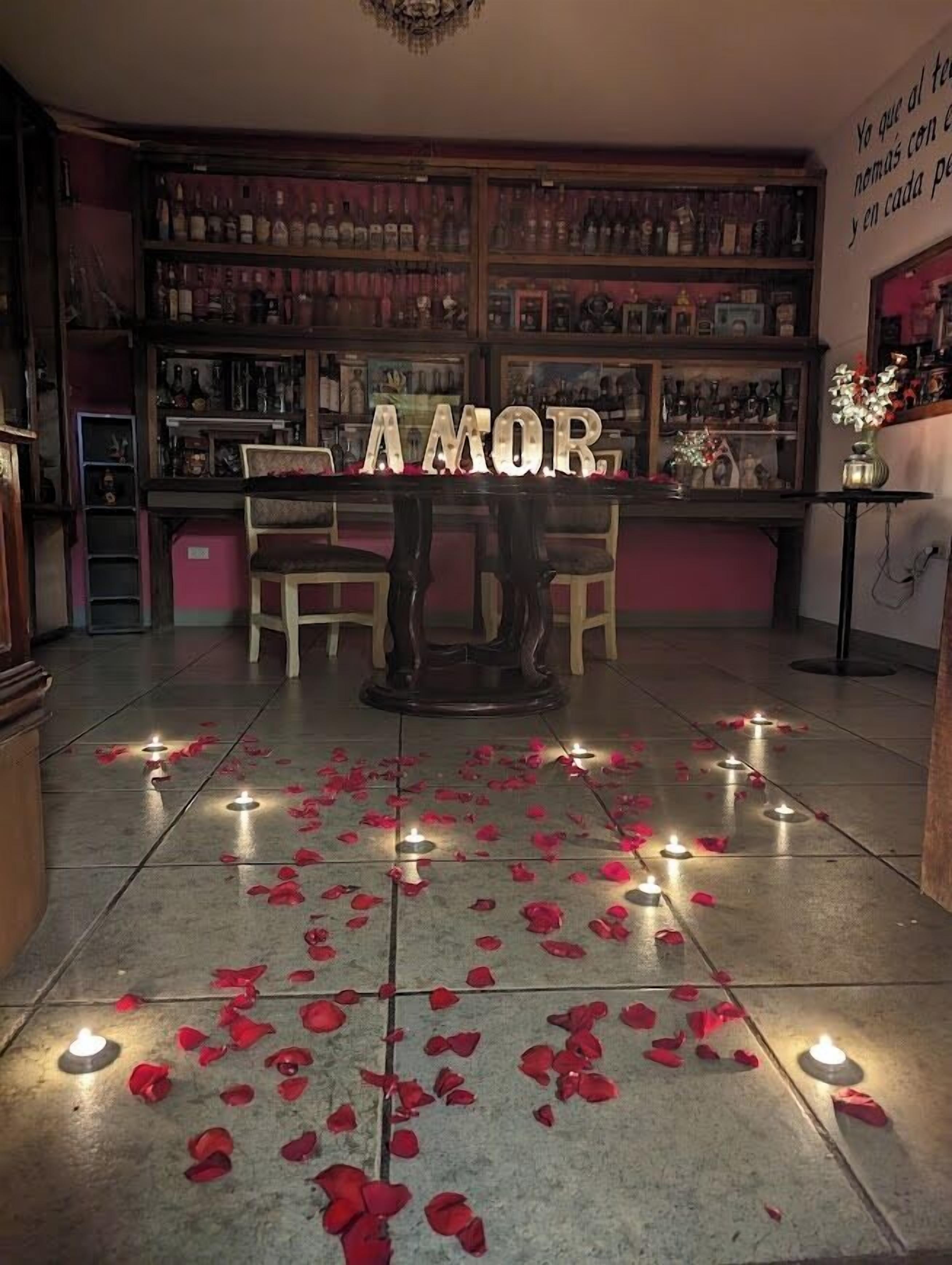 Zona de bar amb un cartell que diu 'AMOR', pètals de rosa al terra i diverses ampolles a les prestatgeries.