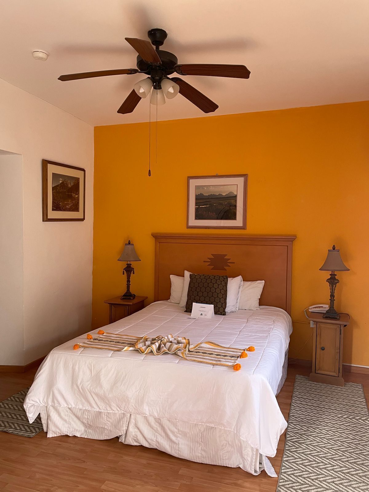 Una habitación de hotel con una cama grande, un ventilador de techo, dos mesitas de noche con lámparas y dos cuadros en las paredes.