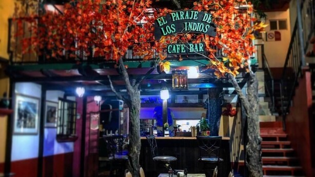 A cafe bar with a sign reading "El Paraje de los Indios Cafe Bar," featuring a rustic interior with wooden tables, chairs, and hanging decorative lights.