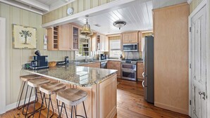 A kitchen with wooden cabinets, a large island with bar stools, a microwave, and a refrigerator. - Beach House on Tidal Creek, Dock, Screen Porch, Easy Walk to Beach, Dog Friendly (Pawleys Island)