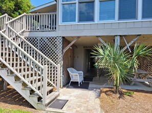 A two-story house with a wooden staircase leading to a balcony, a covered porch area, and a small garden with a palm plant.
