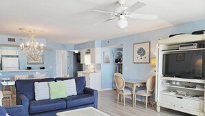 A living room with a blue sofa, a white coffee table, a dining area with chairs, a TV on a stand, and a ceiling fan. - Oceanfront Condo, Top Floor Ocean View, Elevator, Pool, Hot Tub, Beach Access (Pawleys Island)