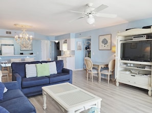 A living room with a blue sofa, a white coffee table, a dining area with chairs, a TV on a stand, and a ceiling fan.