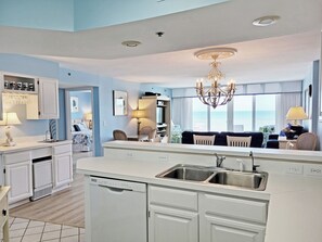A modern kitchen with a central island, a dining area with chairs, a chandelier, and a view of the ocean.