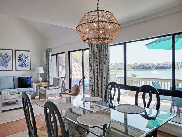 A spacious living area with a glass dining table, black chairs, a rattan chandelier, and a view of a body of water through large windows.