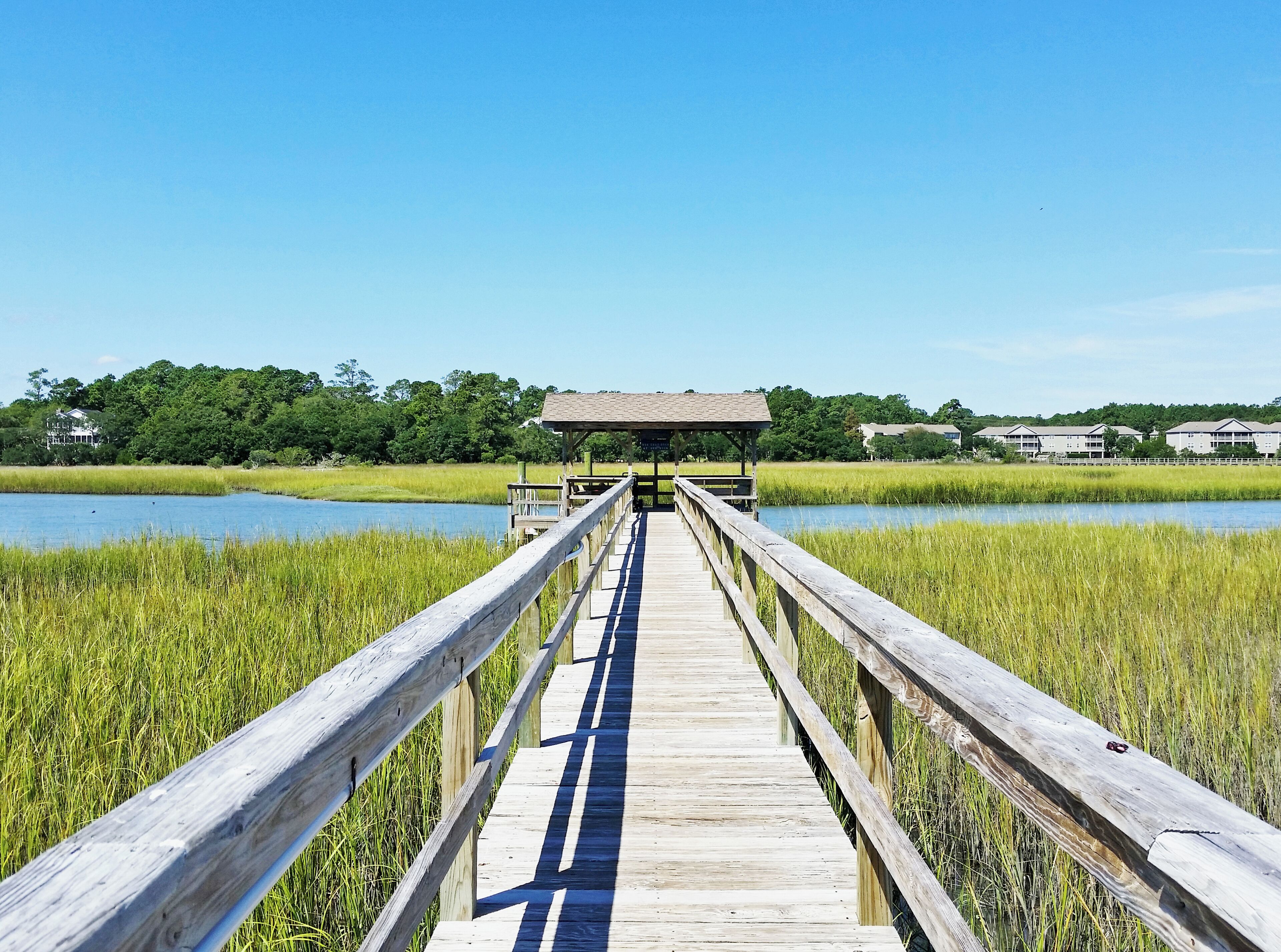 A wooden boardwalk leading to a covered pavilion over a body of water, surrounded by tall grass and trees.