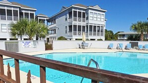 A swimming pool with a wooden fence in the foreground, a lifeguard sign, and a building with balconies in the background.