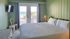 Ein Schlafzimmer mit Bett, Fernseher, Stuhl, Lampe und Blick auf den Strand durch eine Tür.