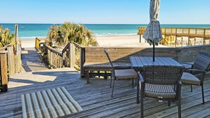 Eine Holzterrasse mit Gartenmöbeln, einem Streifenmuster-Teppich und Blick auf Strand und Meer.