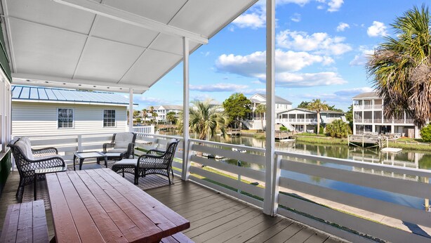 A covered outdoor area with a wooden table and chairs, a view of a lake, and a white building in the background. - Canal Front Beach House, Screened Porch, 2 TV Dens, Dock, Short Block from Beach (Pawleys Island)
