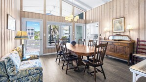 A dining area with a wooden table, chairs, and a chandelier. There is a sofa with patterned cushions and a wooden cabinet with a mirror. - Canal Front Beach House, Screened Porch, 2 TV Dens, Dock, Short Block from Beach (Pawleys Island)