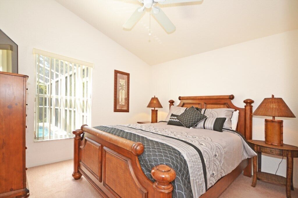 A bedroom with a wooden bed, two bedside lamps, a ceiling fan, a window with blinds, and a wooden dresser.