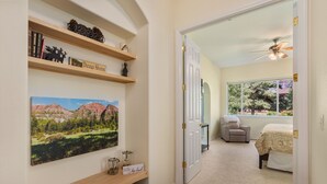 Un pasillo con un cuadro de un paisaje, una estantería llena de libros y objetos decorativos, y una puerta que da a un dormitorio con una cama y una silla.