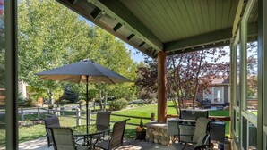 Un patio cubierto con mobiliario exterior, una parrilla y vistas a un jardín ajardinado y edificios cercanos.