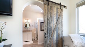 Una habitación con una puerta corredera de estilo granero, un sofá, un televisor montado en la pared y vistas a un baño con tocador y espejo.