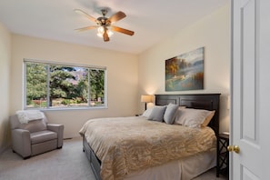 A bedroom with a bed, a chair, a ceiling fan, a window with a view of trees, and a painting on the wall.