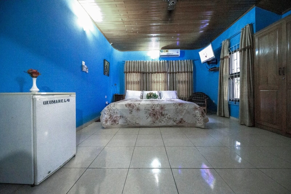 A bedroom with a bed, a refrigerator, a window with curtains, and a television on the wall.
