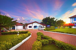 A hotel with a paved driveway leading to a white building with a red roof. There are neatly trimmed bushes and a lawn in the foreground. - Okumah Executive Lodge & Conference Centre (Kintampo)
