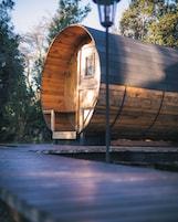 Una estructura cilíndrica de madera con una puerta y ventana, ubicada al aire libre con árboles en el fondo.