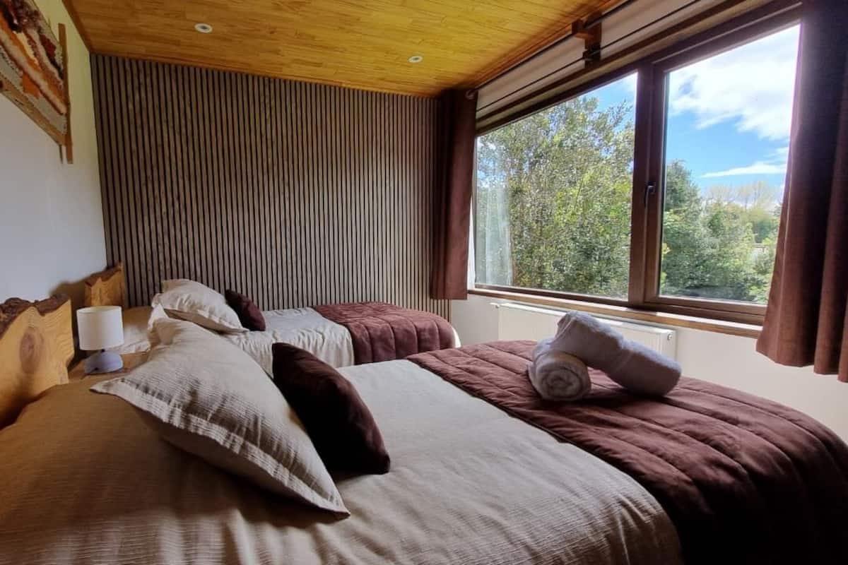 Een slaapkamer met twee bedden, een houten hoofdbord, een raam met uitzicht op bomen en een plafond met houten balken.