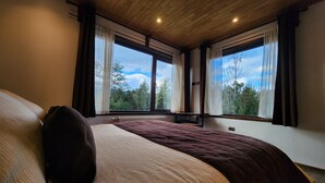 Un dormitorio con una cama grande, dos ventanas con cortinas, techo de madera y vistas a árboles.