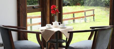 Un rincón con asientos, una mesa dispuesta para dos, un jarrón con flores rojas y vistas a un espacio verde a través de amplias ventanas.