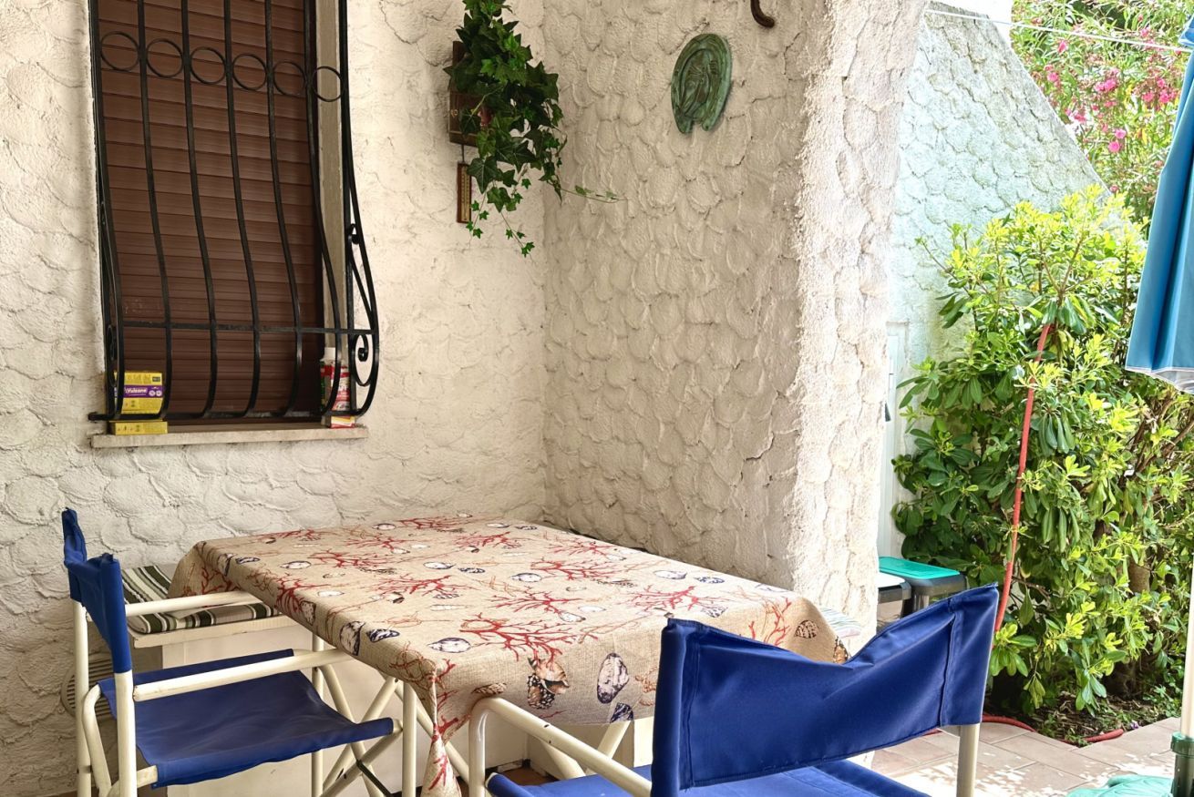 A small outdoor seating area with a table and chairs, a window with a metal grill, and a wall with a textured surface and a green plant.