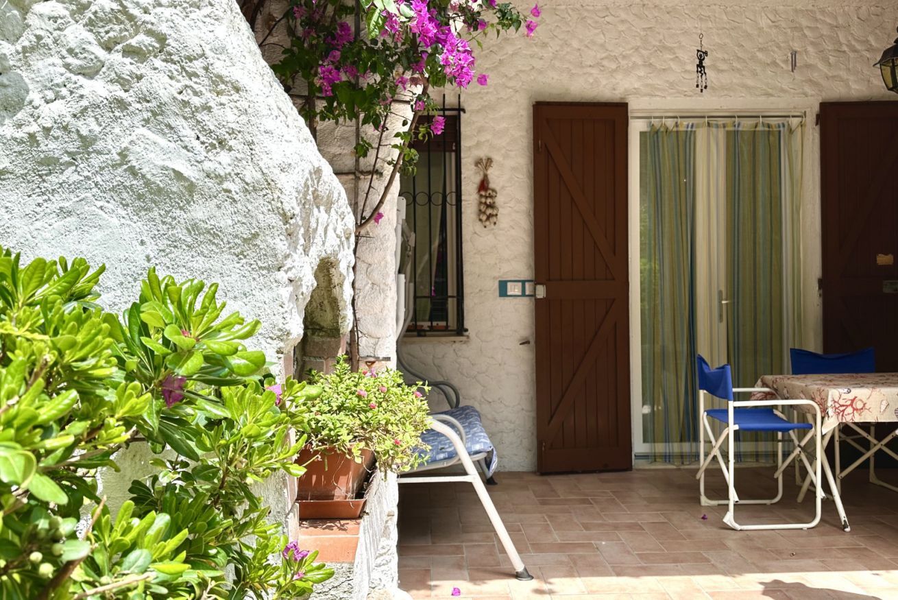 Un edificio in pietra con una porta, una finestra con cancello metallico, un tavolo con sedie e piante in vaso.