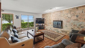 A living room with a stone wall, a flat-screen TV, a fireplace, and a large window overlooking greenery. - EZ Access Lake House (Quinlan)