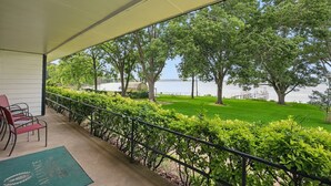 A covered balcony with a view of a lake, green lawn, and trees. - EZ Access Lake House (Quinlan)