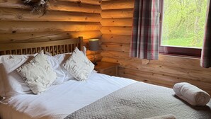 Een houten kamer met een bed, kussens, een nachtkastje, een lamp, een raam met gordijnen en een krans aan de muur.
