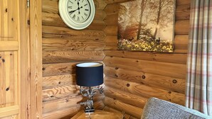 Een knus houten interieur met een klok, lamp, schilderij en een ruitjesgordijn.