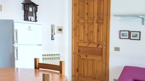 Una habitación con puerta de madera, un refrigerador blanco, una mesa de madera, un sofá morado y una estantería de pared.