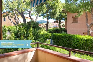 A balcony with a metal railing, surrounded by lush greenery and a view of a building with a red roof.