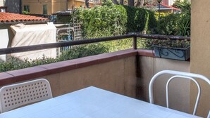 Un balcón con una mesa y sillas blancas, barandilla de metal y vista a edificios y follaje.