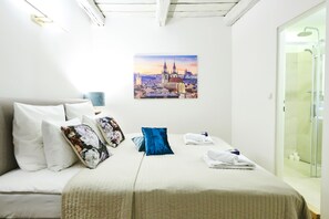A hotel room with a bed, pillows, a neatly made bed, a glass shower, and a cityscape painting on the wall.