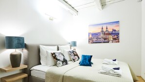 Una camera d'albergo ordinata con un letto, cuscini, un comodino con una lampada, un quadro appeso al muro e un asciugamano piegato sul letto.
