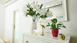Un mobile bagno bianco con uno specchio, un vaso di fiori bianchi, una piccola candela e due piante in vaso.