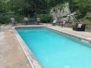 A rectangular swimming pool with a tiled edge, surrounded by a concrete deck. There are four lounge chairs and a small table with a canopy overhead. A large rock with a pipe protruding from it is visible to the right of the pool.