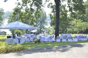 An outdoor event setup with tables and chairs arranged on a grassy area, a blue canopy tent, and a lake in the background.