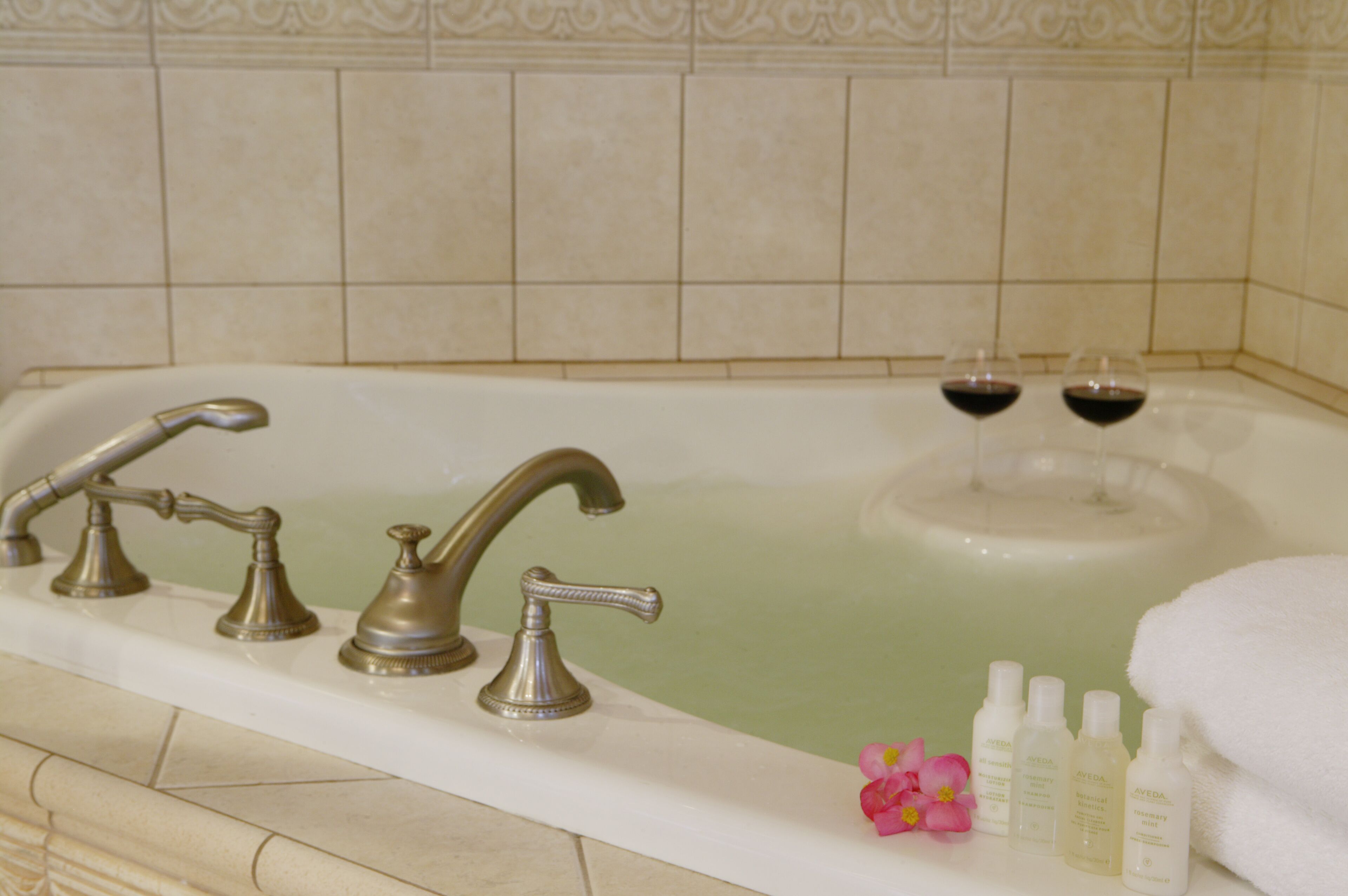 A bathtub with two wine glasses, a towel, and three bottles of Aveda products.