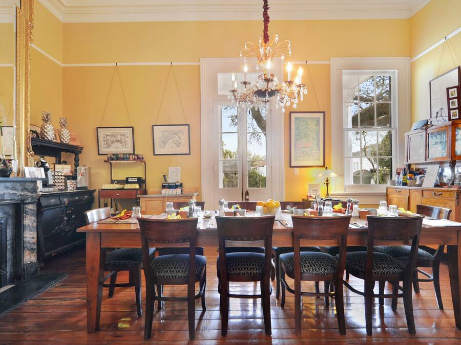 A dining room with a wooden table set for six, surrounded by patterned chairs. A chandelier hangs from the ceiling, and the room features a large window with a view of trees.