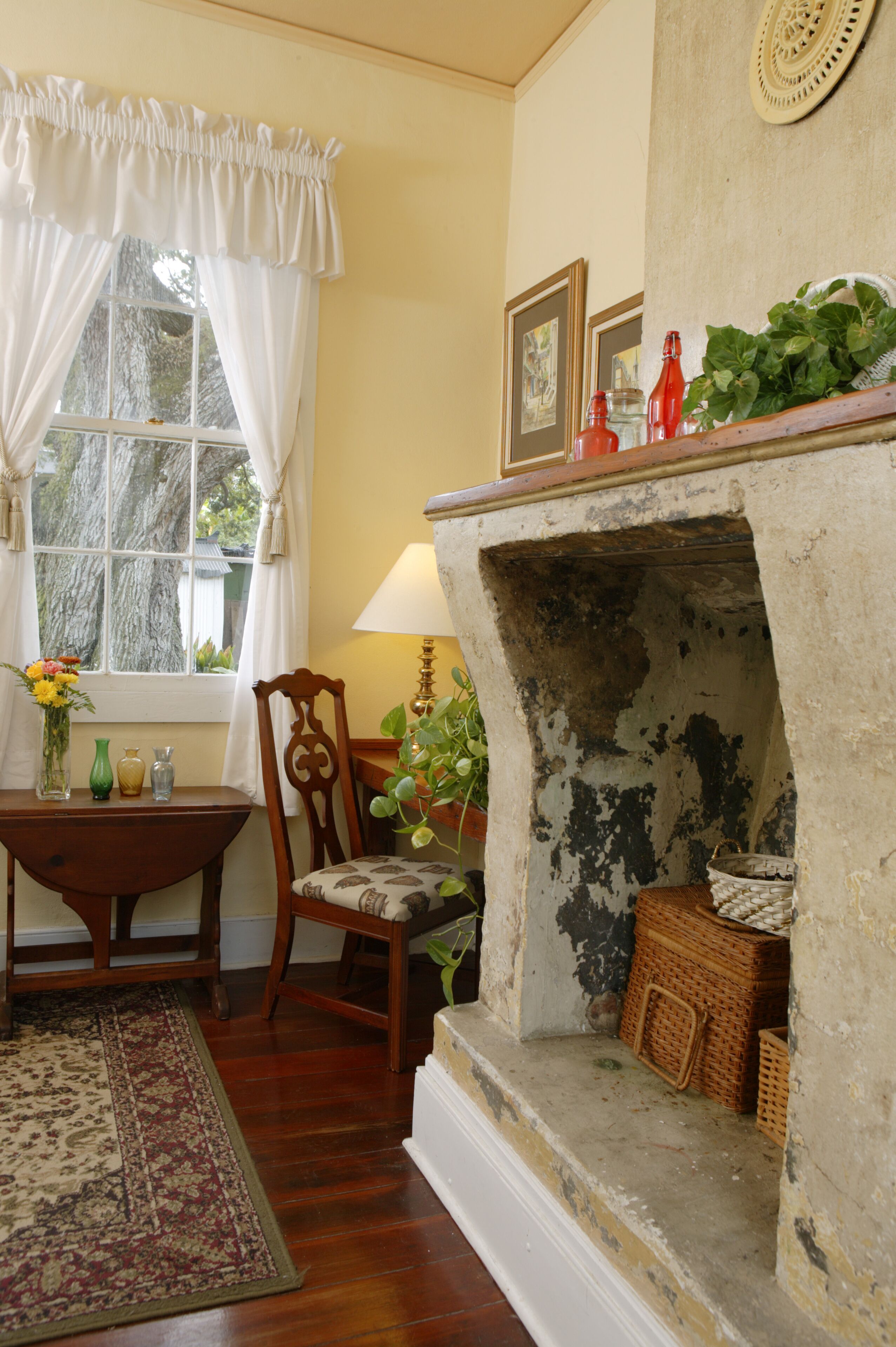 A cozy room with a fireplace, a wooden chair, a table with a vase of flowers, and a window with curtains.