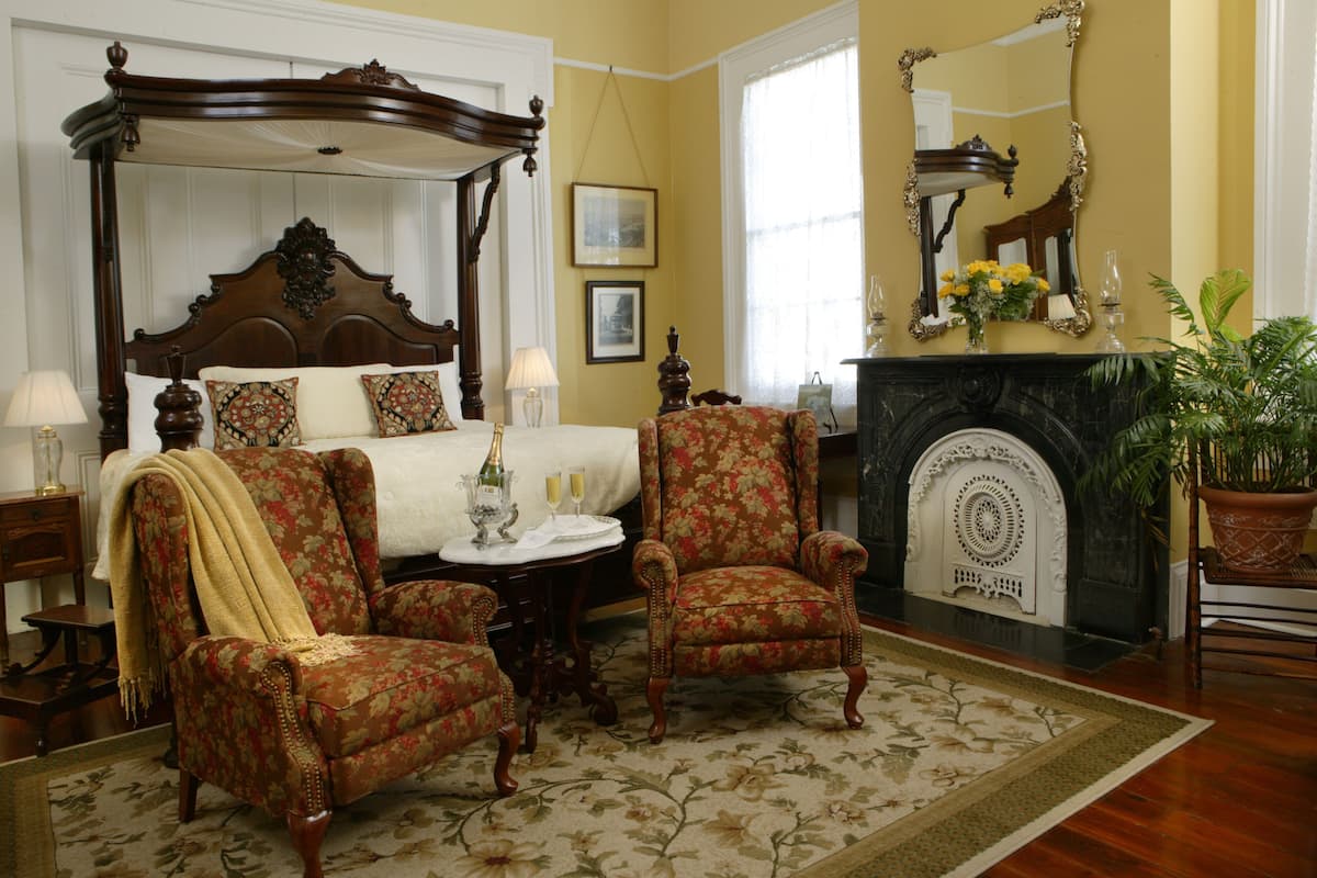 Quarto com cama de dossel, duas poltronas, lareira e um espelho grande.