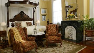 A bedroom with a four-poster bed, two armchairs, a fireplace, and a large mirror.