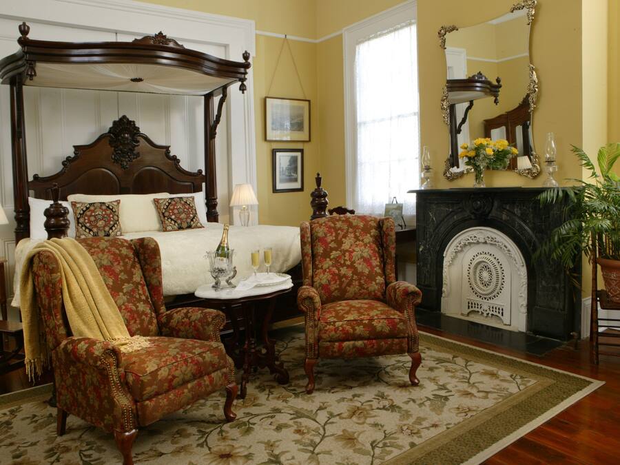 A bedroom with a four-poster bed, two armchairs, a fireplace, and a large mirror.